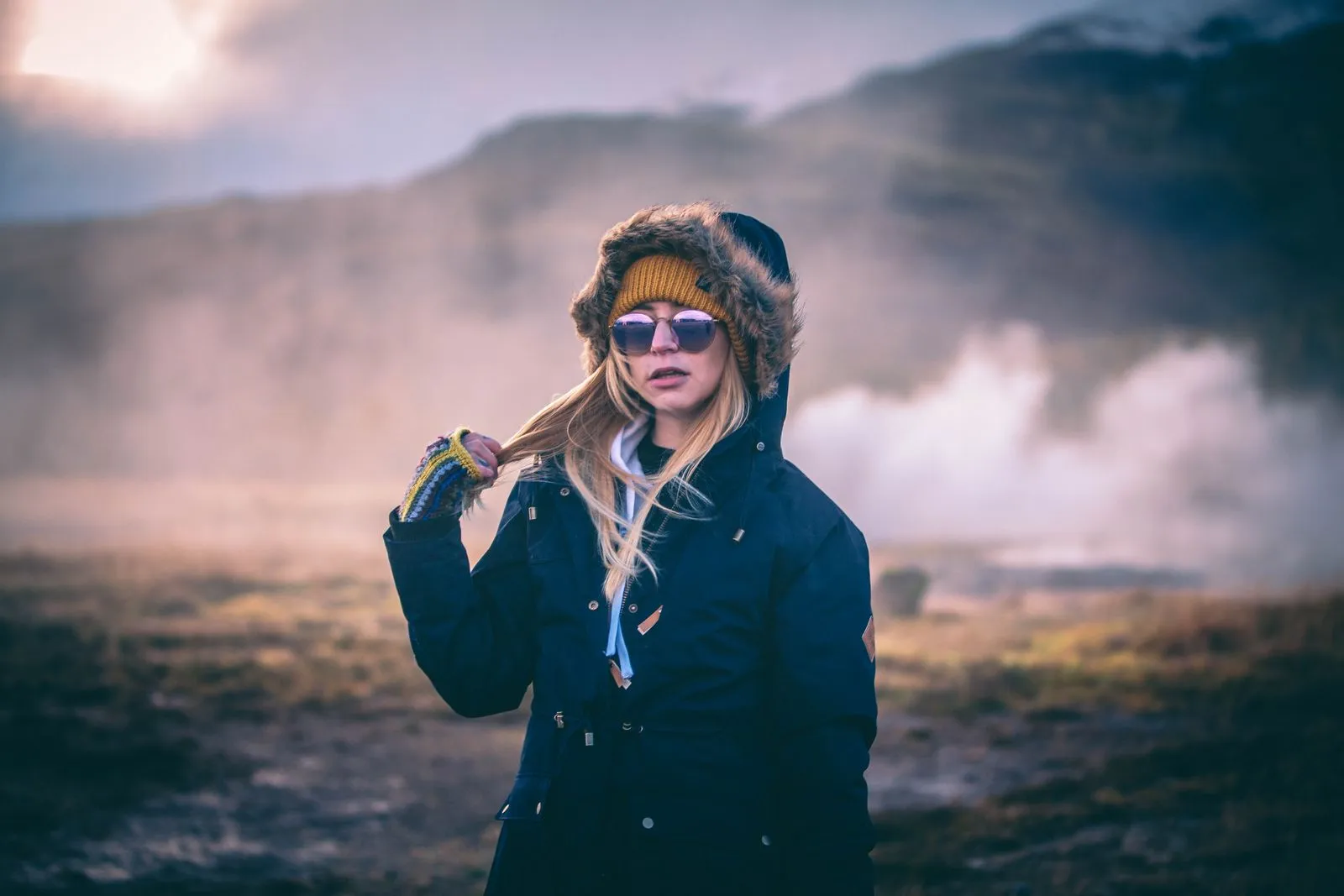 A fashionable woman in a parka stands in a scenic, foggy Icelandic landscape, showcasing winter style.